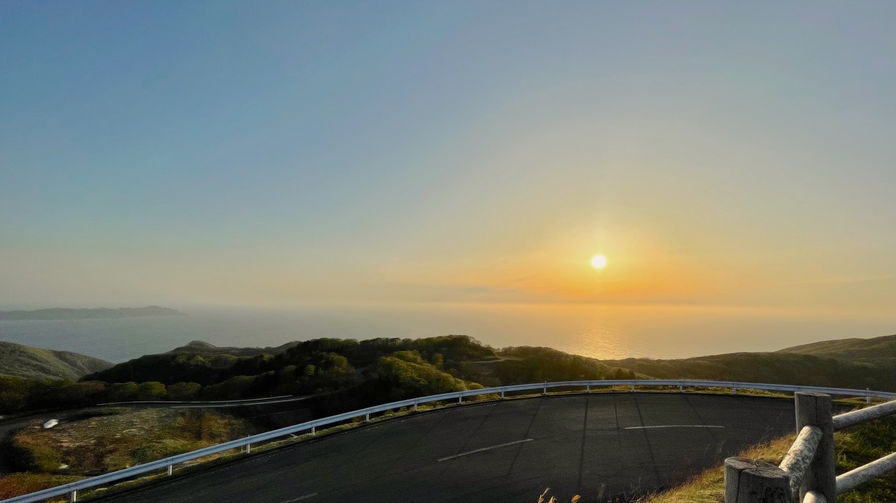 The sun low over the sea beyond a winding coastal road and green hills.