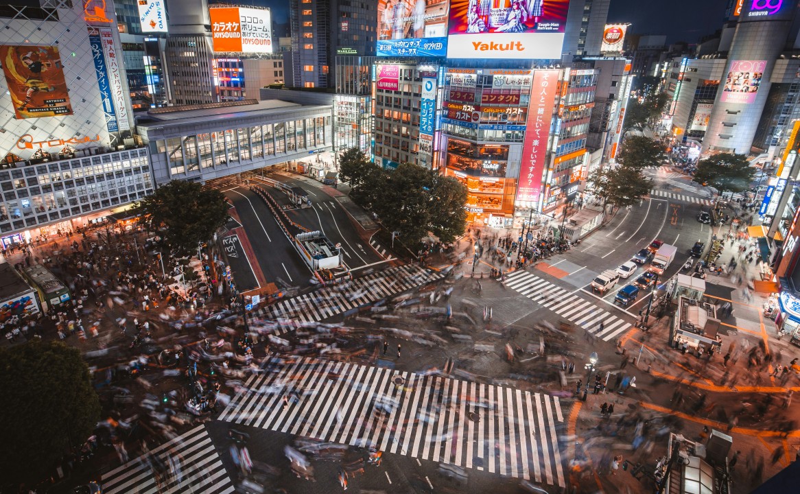 Crowds crossing Shibuya's intersecting pedestrian crossings beneath illuminated buildings at night.
